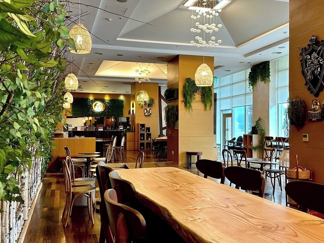 A spacious indoor area with wooden tables and chairs, hanging pendant lights, and large windows allowing natural light.