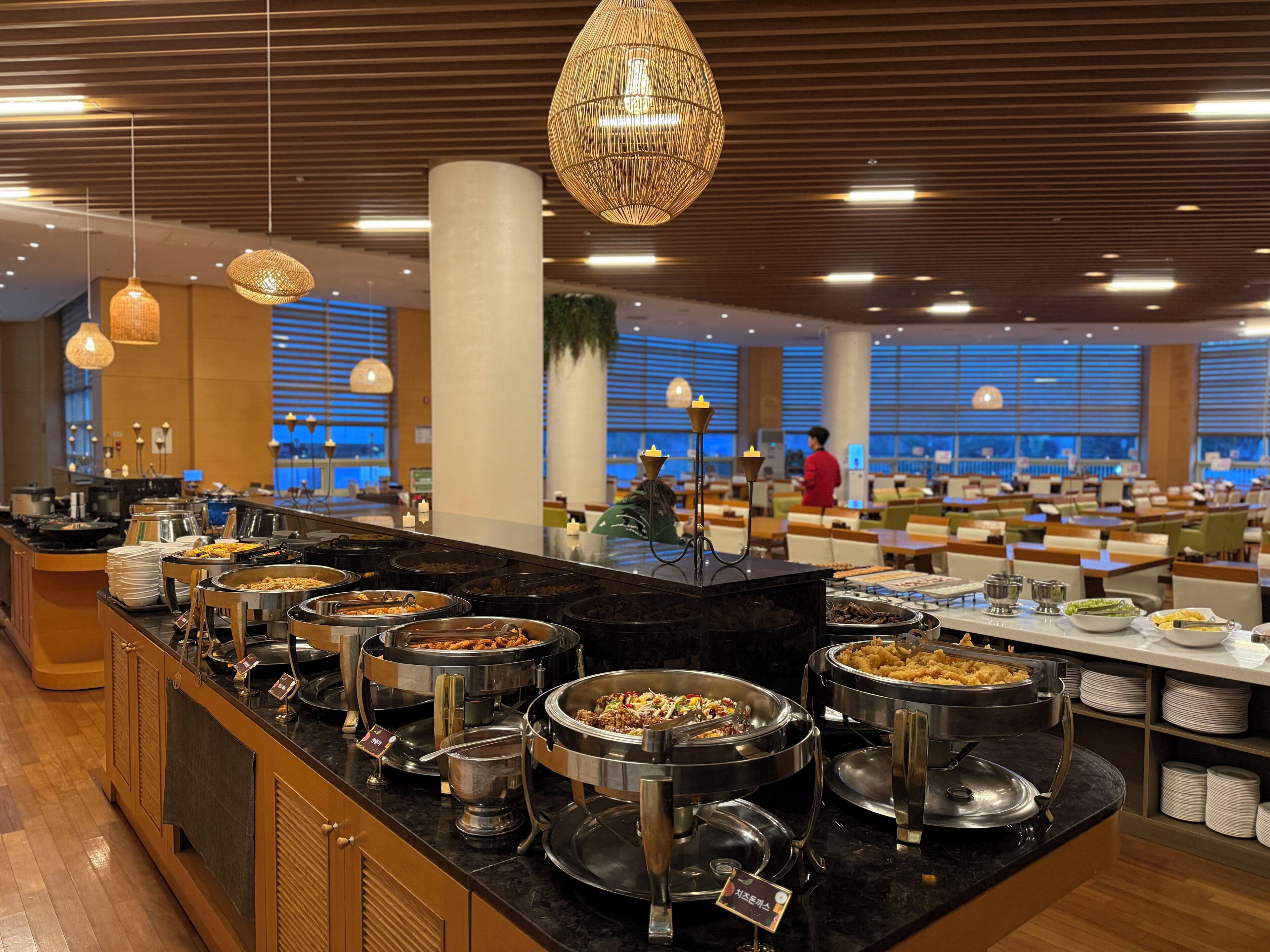 A buffet area with multiple food stations, a seating area with tables and chairs, and a view of the outdoors through large windows.