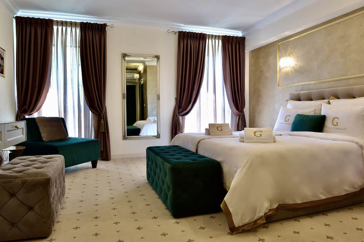 A hotel room with a large bed, a green ottoman, a tufted chair, and a mirror.