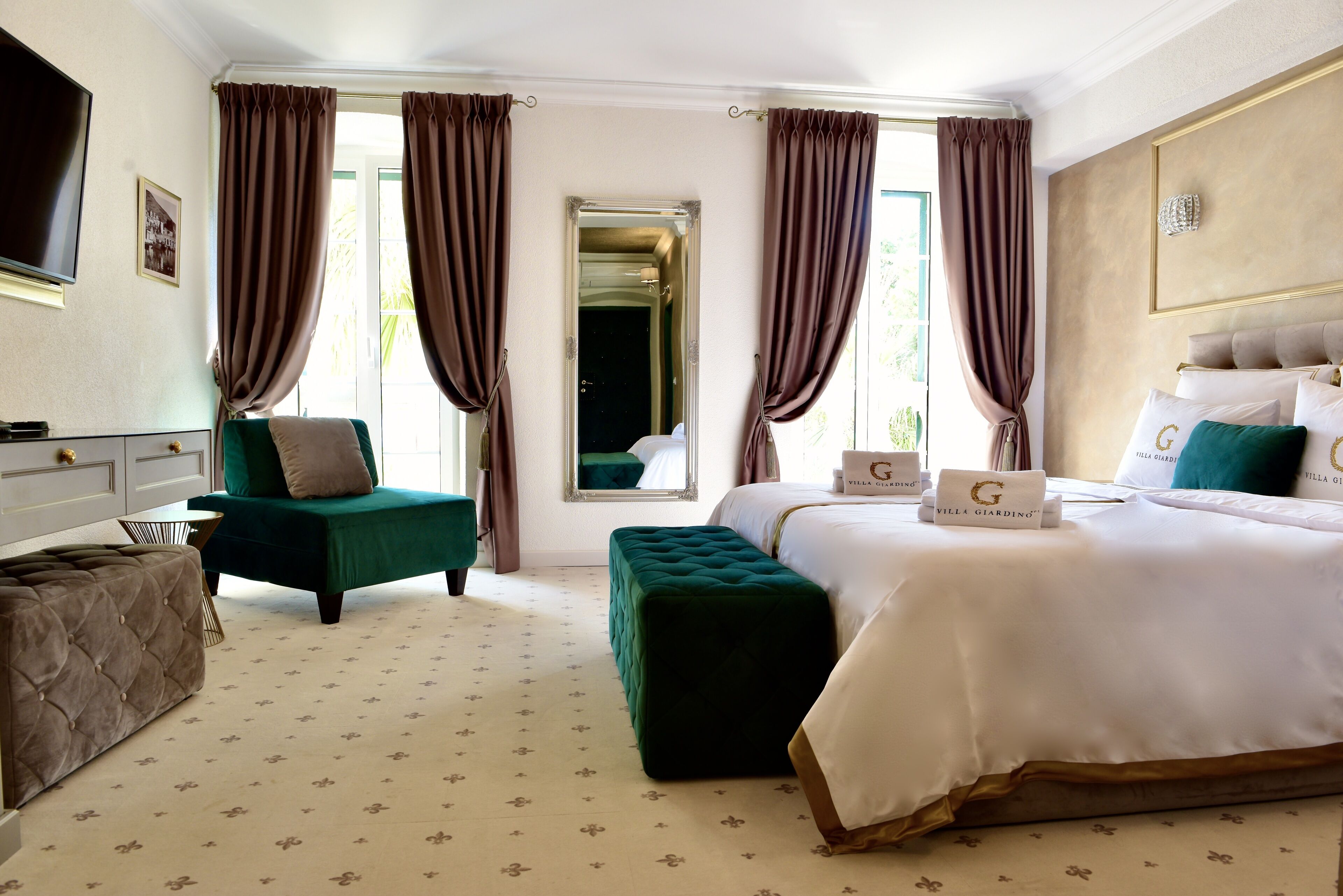 A hotel room with a large bed, a green armchair, a matching ottoman, a flat-screen TV, and a view of the outdoors through a window with curtains.