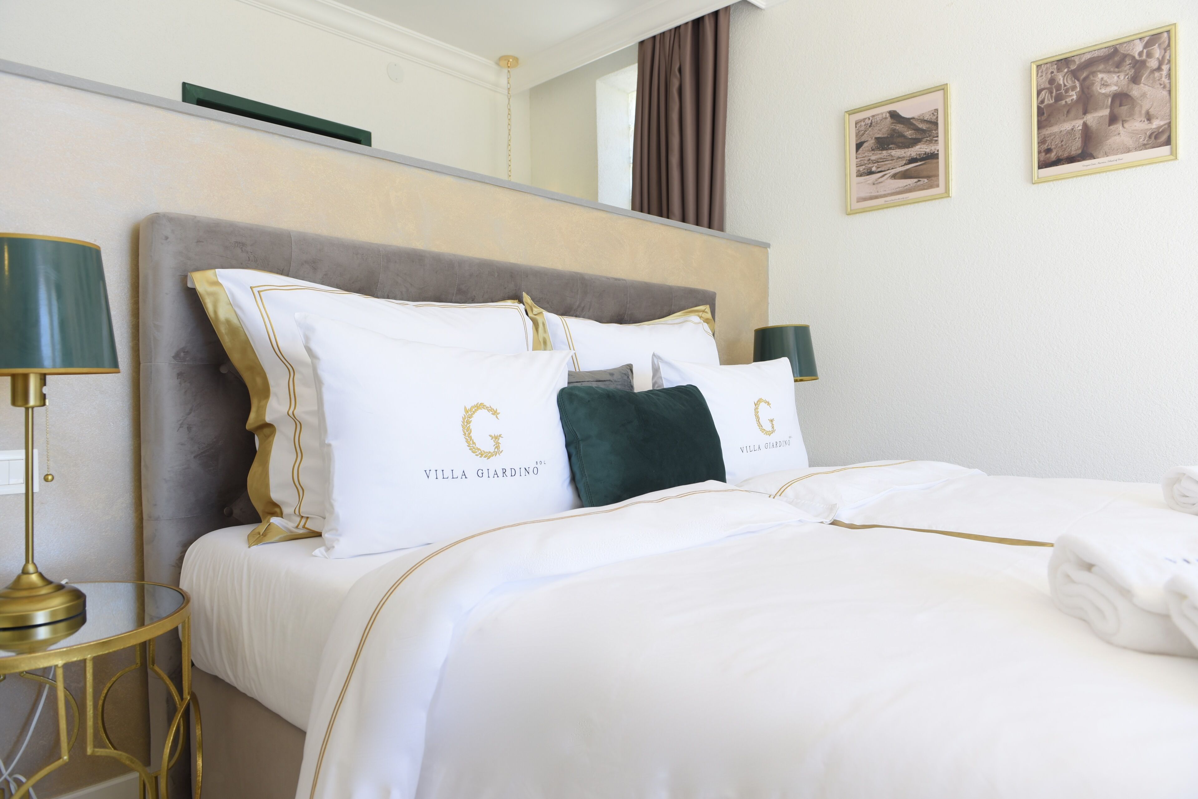 A hotel room with a large bed, two pillows with 'Villa Giardino' branding, a gold-framed lamp, and a framed picture on the wall.