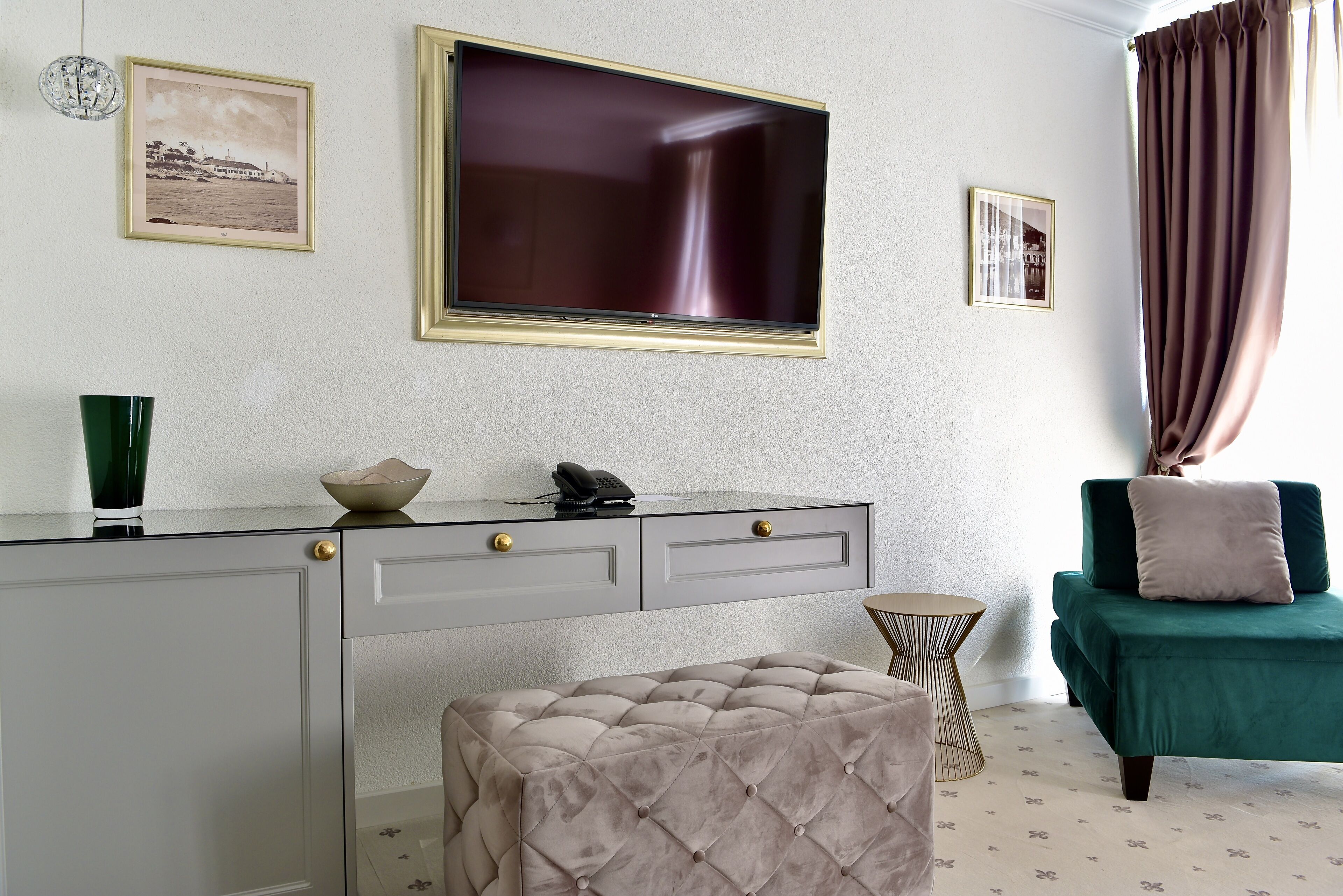 A room with a TV mounted on the wall, a desk with drawers, a cushioned ottoman, a green armchair, and a small side table.
