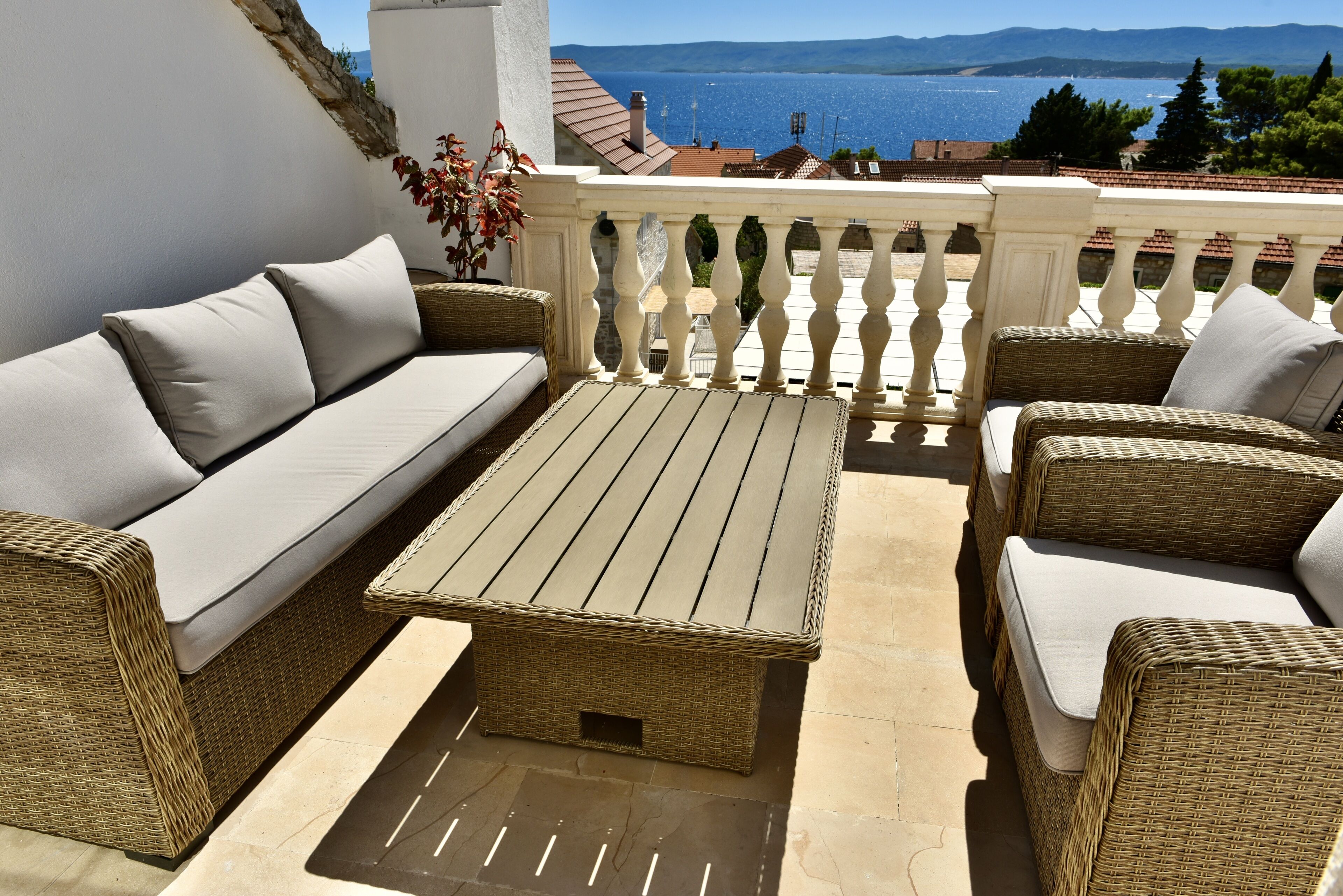 A balcony with wicker furniture, including a sofa and a table, overlooking a scenic view of the sea and mountains.