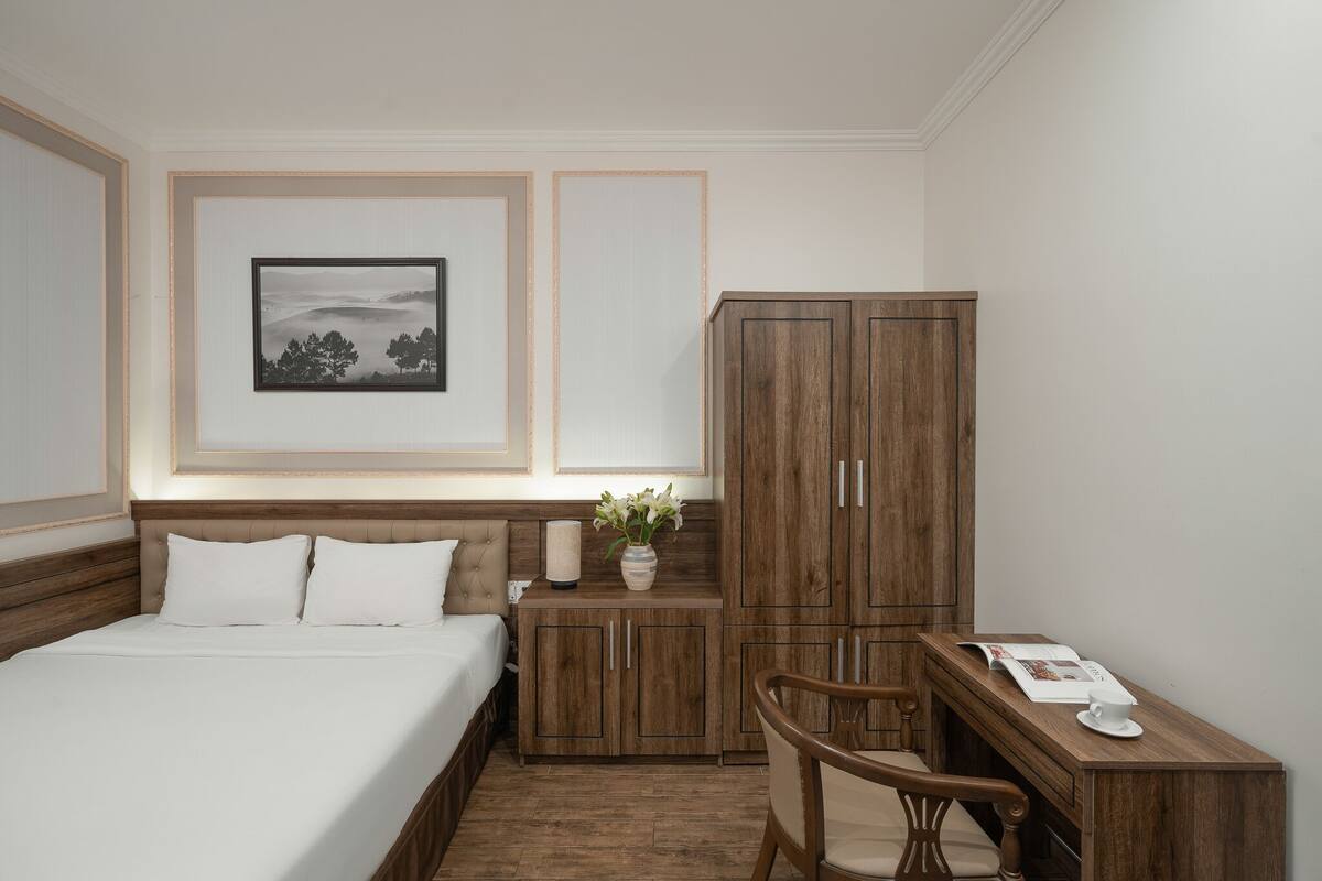 A hotel room with a bed, a wooden wardrobe, a small table with a chair, and a framed picture on the wall.