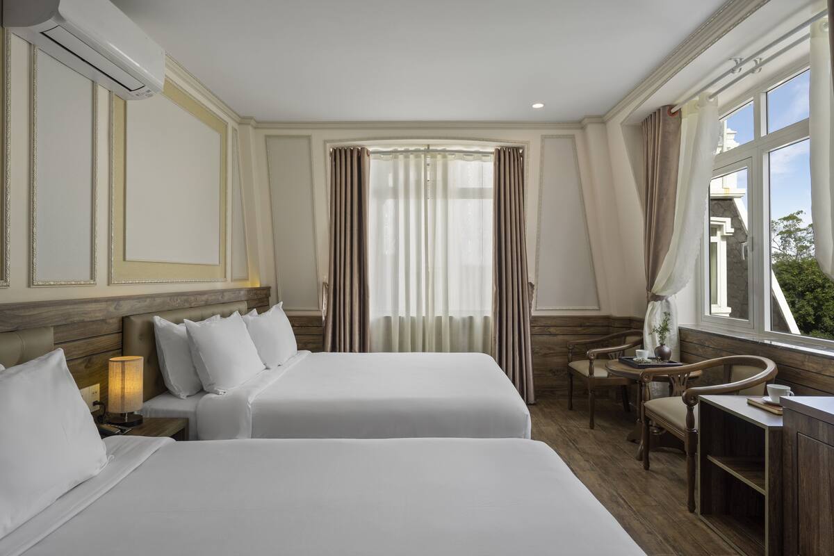 A hotel room with two beds, a wooden headboard, a small table with a lamp, and a view of the outdoors through large windows.