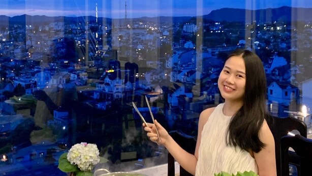 A person holding chopsticks and a plate of food, with a table of various dishes and a hot pot in the foreground. - New Life Dalat Hotel (Xuan Huong - Da Lat)