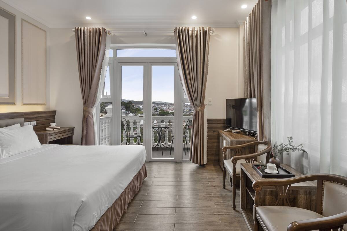A hotel room with a large bed, a TV, a balcony with curtains, and a dining area with chairs and a table.