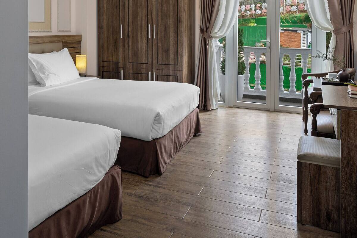 A hotel room with two beds, a wooden wardrobe, a desk with a chair, and a balcony with curtains.