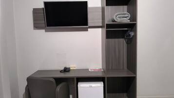 Un espacio de oficina compacto con un escritorio, dos sillas, un refrigerador pequeño, un televisor de pantalla plana montado en la pared y una estantería con toallas dobladas.