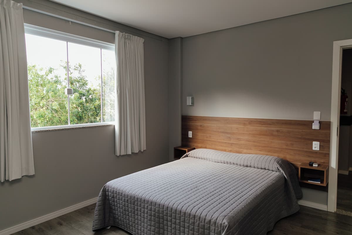 Une chambre à coucher comprenant un lit, une table de chevet, une tête de lit en bois, une fenêtre avec des rideaux et une vue sur les arbres à l’extérieur.