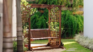 Een houten pergola met een bankje in een tuin met een grindpad, omringd door groen en een wit gebouw op de achtergrond.