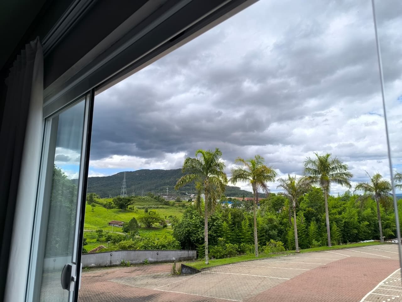 A view from a window with a view of a parking lot, palm trees, and a green landscape with hills in the background.