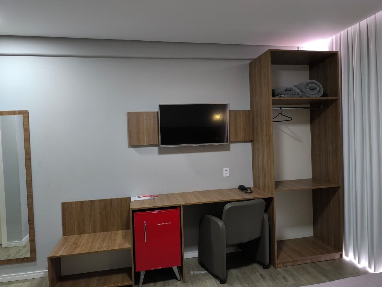 Habitación compacta con escritorio de madera, un mini refrigerador rojo, televisor montado en la pared y armario con espacio para colgar ropa.