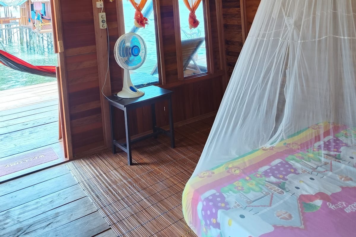 A wooden cabin with a thatched roof, a bed with a colorful bedspread, a fan, and a view of a dock and water.