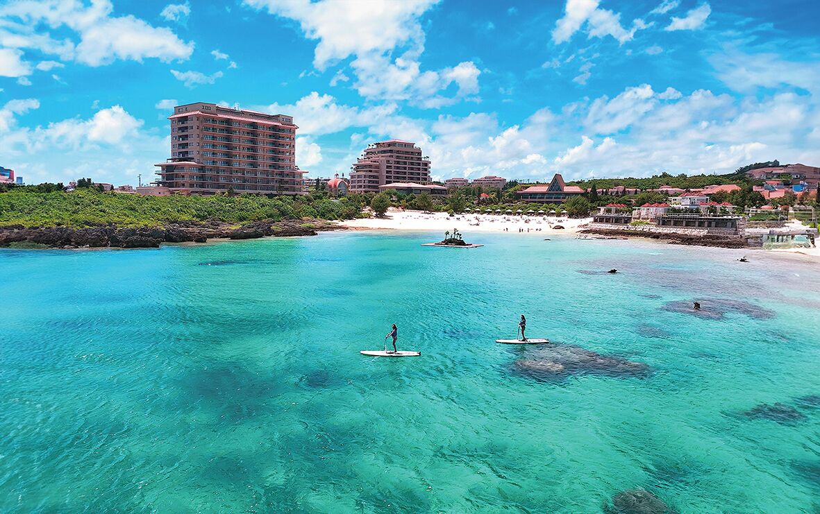A coastal area with clear turquoise water, paddleboarders, and buildings in the background.