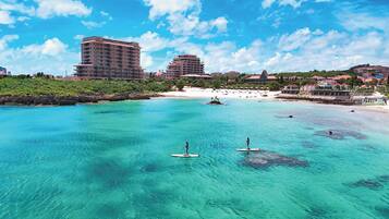 A coastal area with clear turquoise water, paddleboarders, and buildings in the background.
