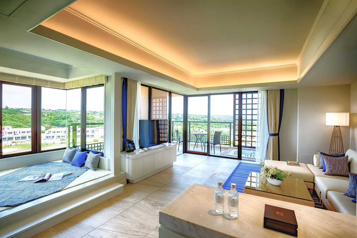 A spacious hotel room with a large window offering a city view, a comfortable seating area with a sofa and a coffee table, and a flat-screen TV on a white console.