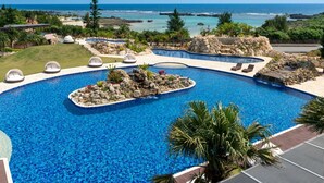 A large, clear blue pool with multiple sections and surrounding greenery, overlooking a coastal area. - Hotel Shigira Mirage (Miyakojima)