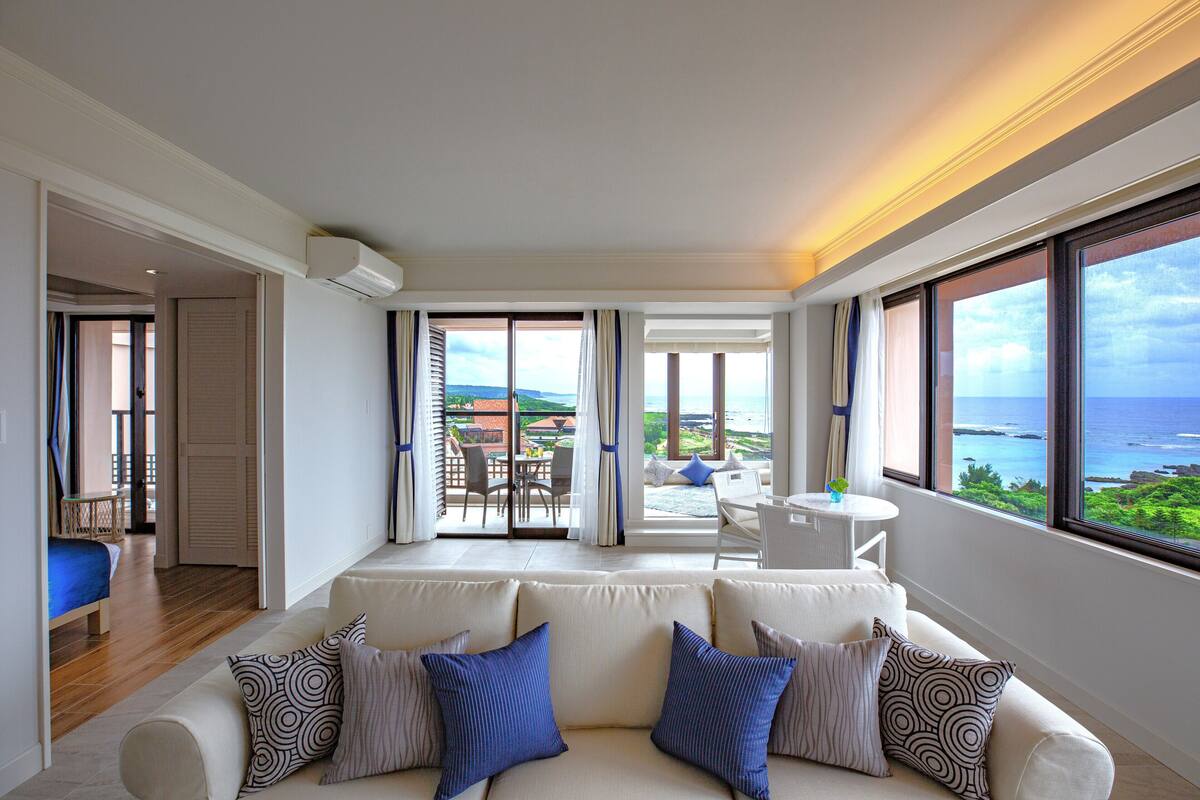 A modern living room with a sofa, a dining area, and a view of the sea.