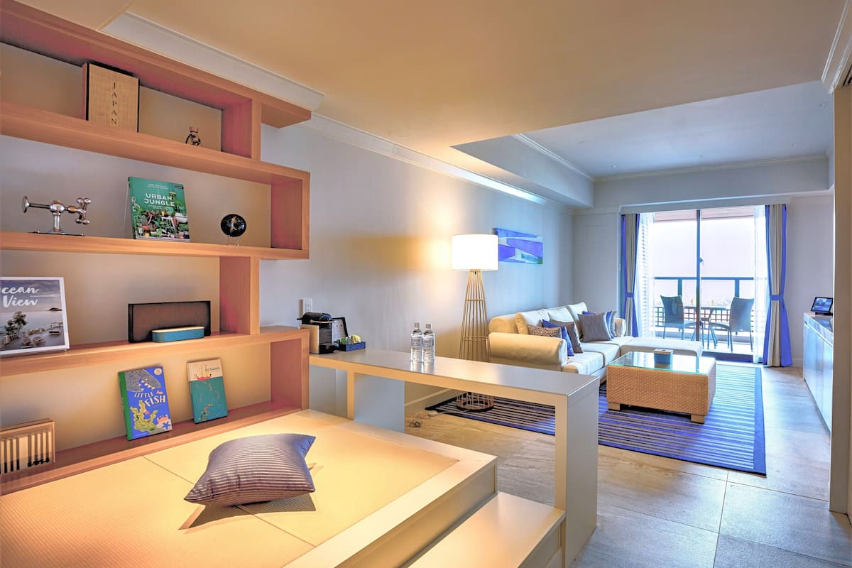 A modern hotel room with a sofa, a coffee table, a bookshelf, and a view of the outside balcony.