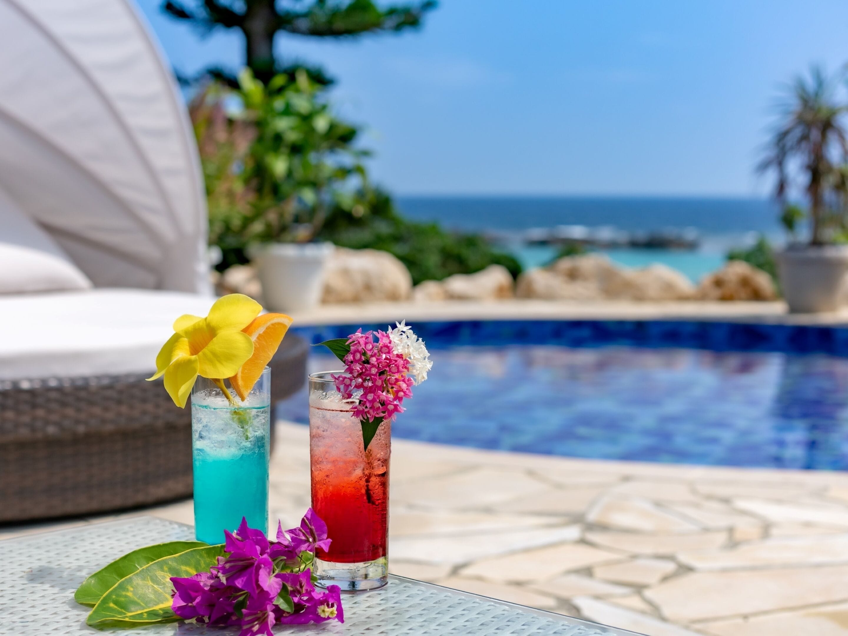 A poolside setting with two drinks, one with a yellow flower and the other with pink flowers, and a view of the sea and rocks in the background.