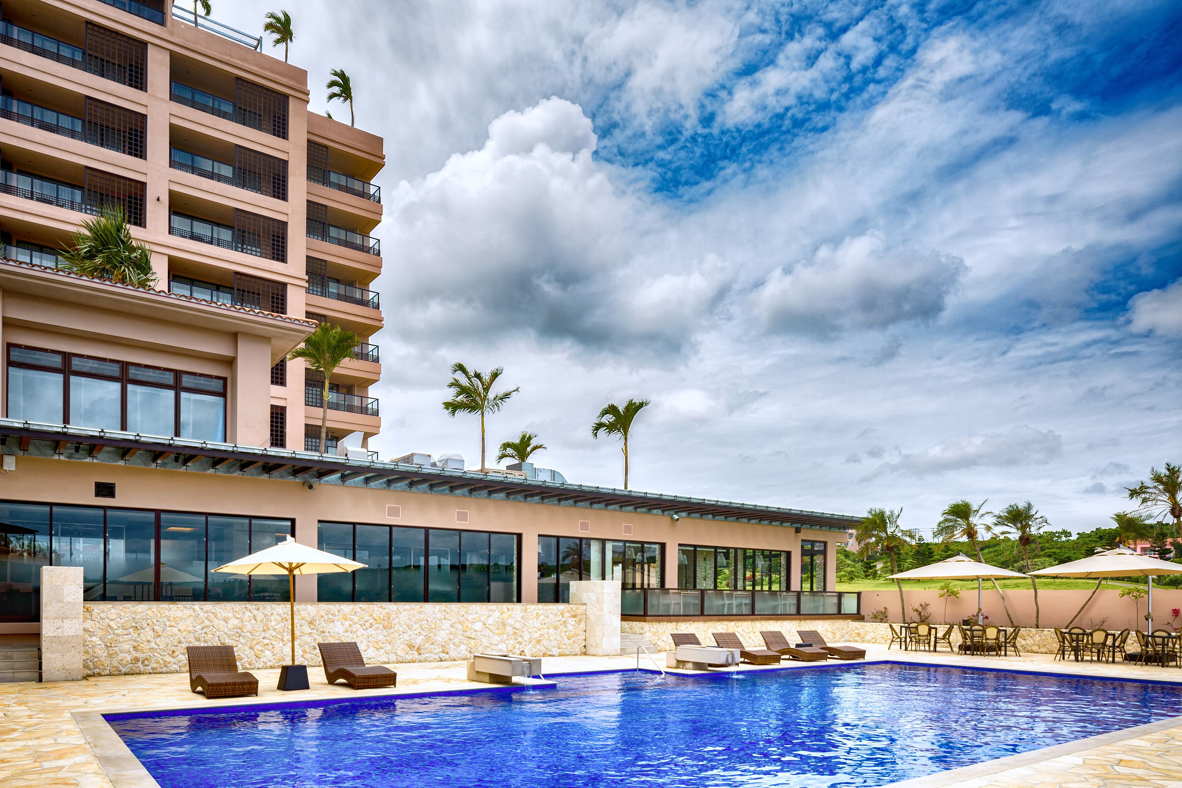 A swimming pool with lounge chairs and umbrellas, a stone wall, and a building with multiple floors and balconies.