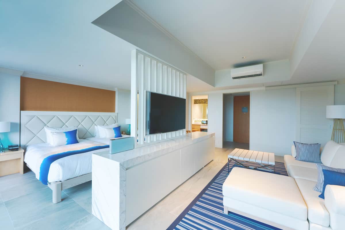 A modern hotel room with a large bed, a TV mounted on a white console, a seating area with a sofa and armchairs, and a small kitchenette.