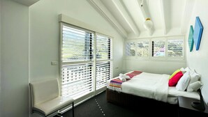 Une chambre d’hôtel moderne avec une grande fenêtre, un lit agrémenté de coussins colorés et un petit bureau avec une lampe.