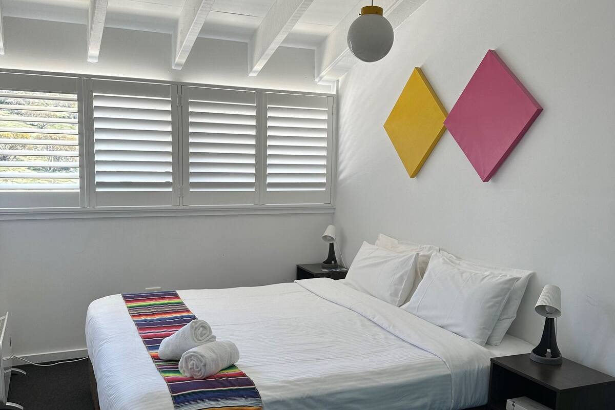 Ein ordentlich bezogenes Bett mit weißen Leinen und einer farbenfrohen, gestreiften Decke, ein Nachttisch mit Lampe sowie zwei Wandleuchten.