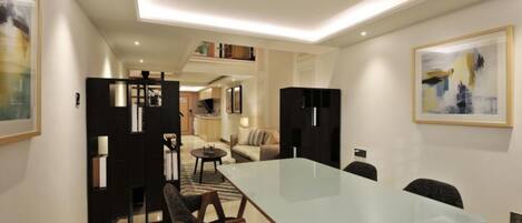 A modern hotel room with a glass dining table, a sofa, a small coffee table, and a bookshelf.