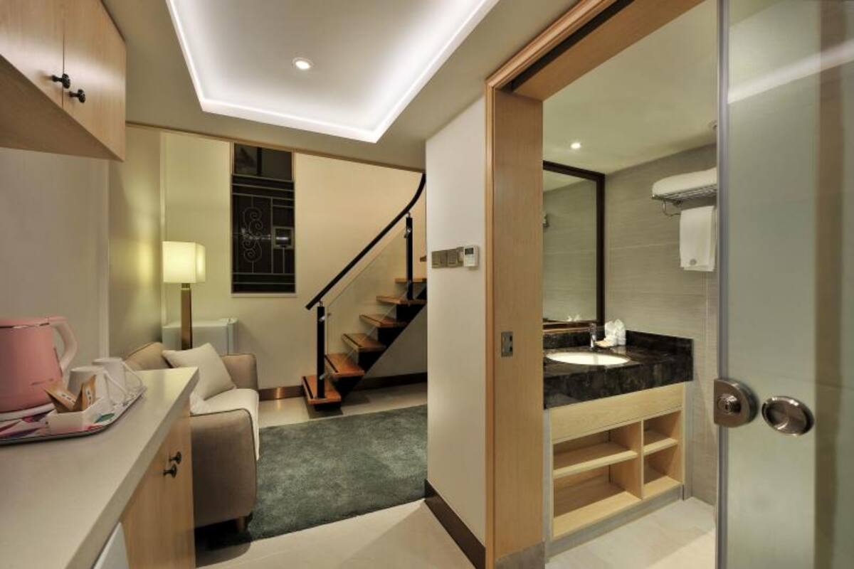 A modern hotel room with a sofa, a coffee table, a staircase, and a bathroom with a glass shower enclosure.