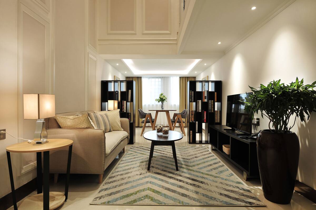 A modern hotel room with a sofa, a small round table, a TV, a bookshelf, and a large potted plant.