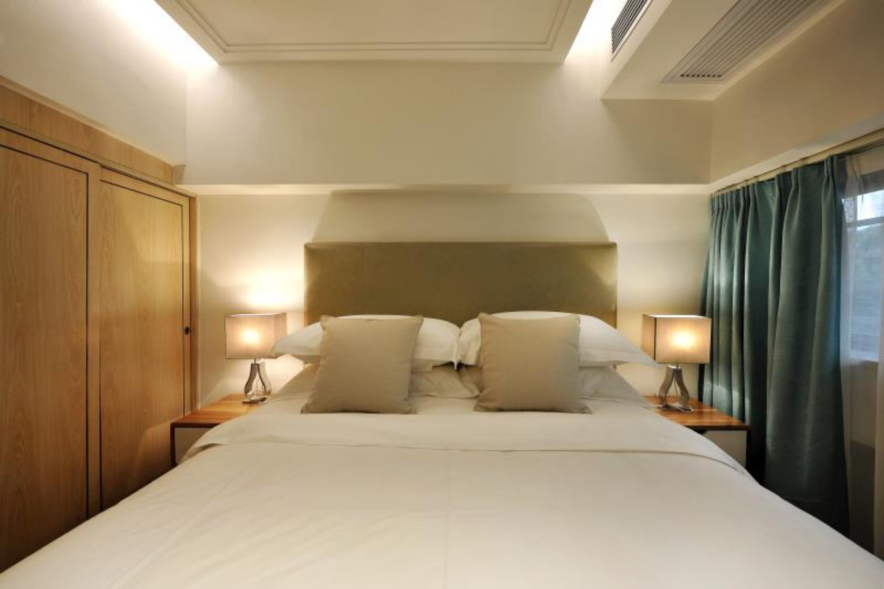 A hotel room with a large bed, two bedside lamps, a wooden wardrobe, and a view of the outside through a window with curtains.