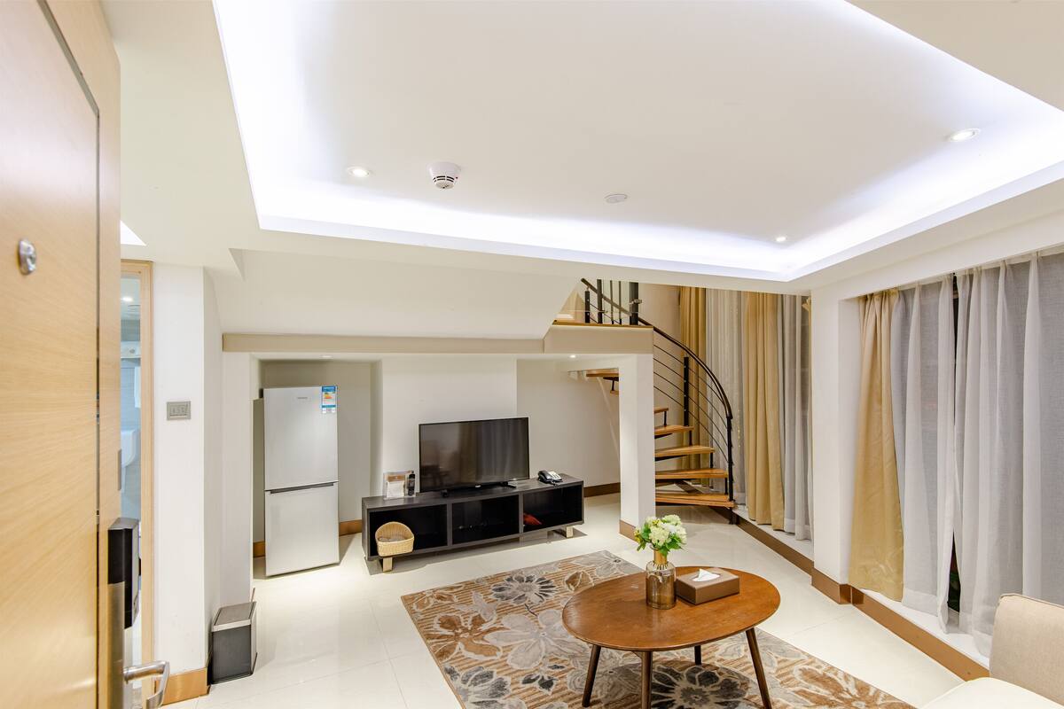 A modern living room with a flat-screen TV, a dining table, and a staircase leading to an upper level.
