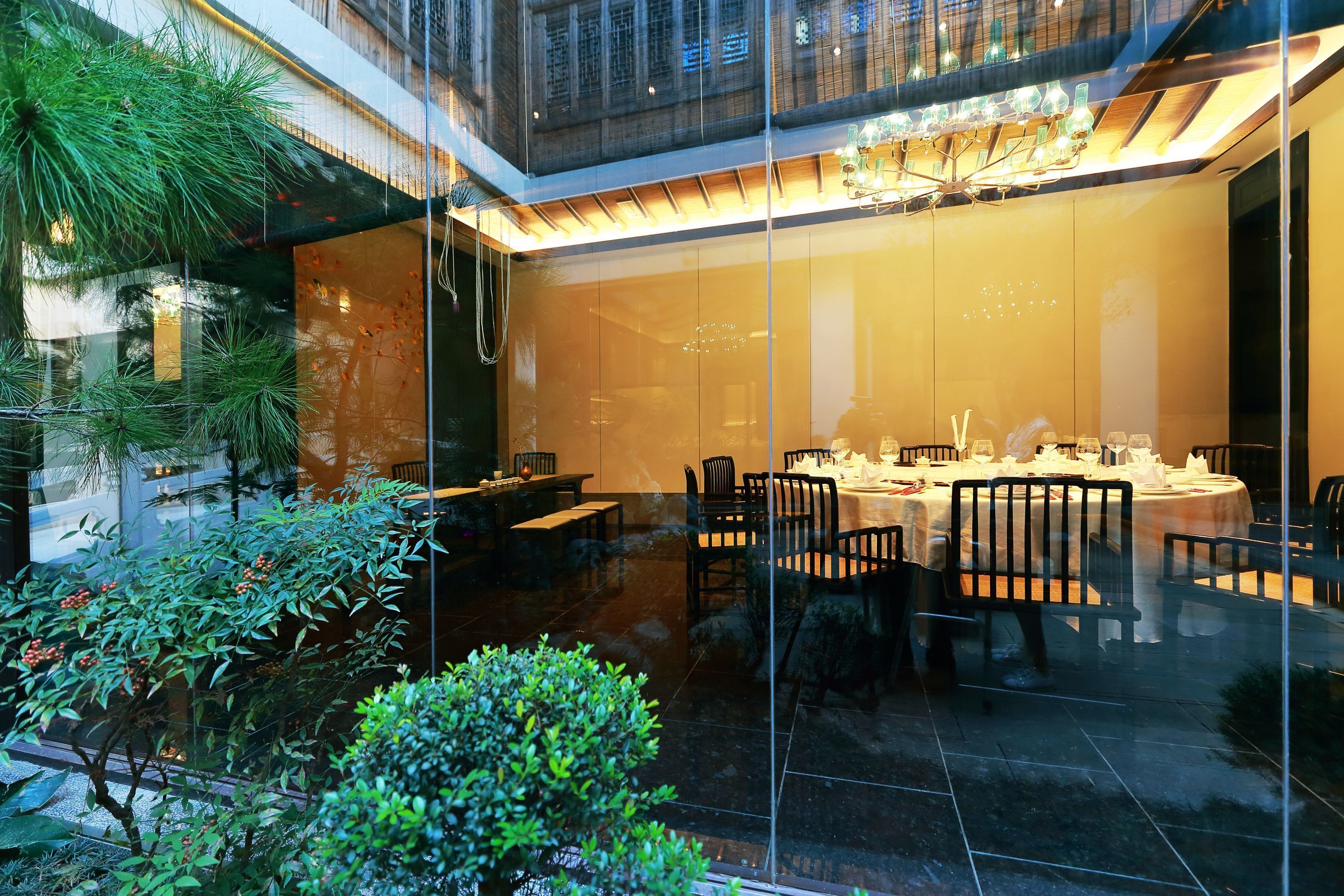 A modern dining area with glass walls, a chandelier, and indoor plants.