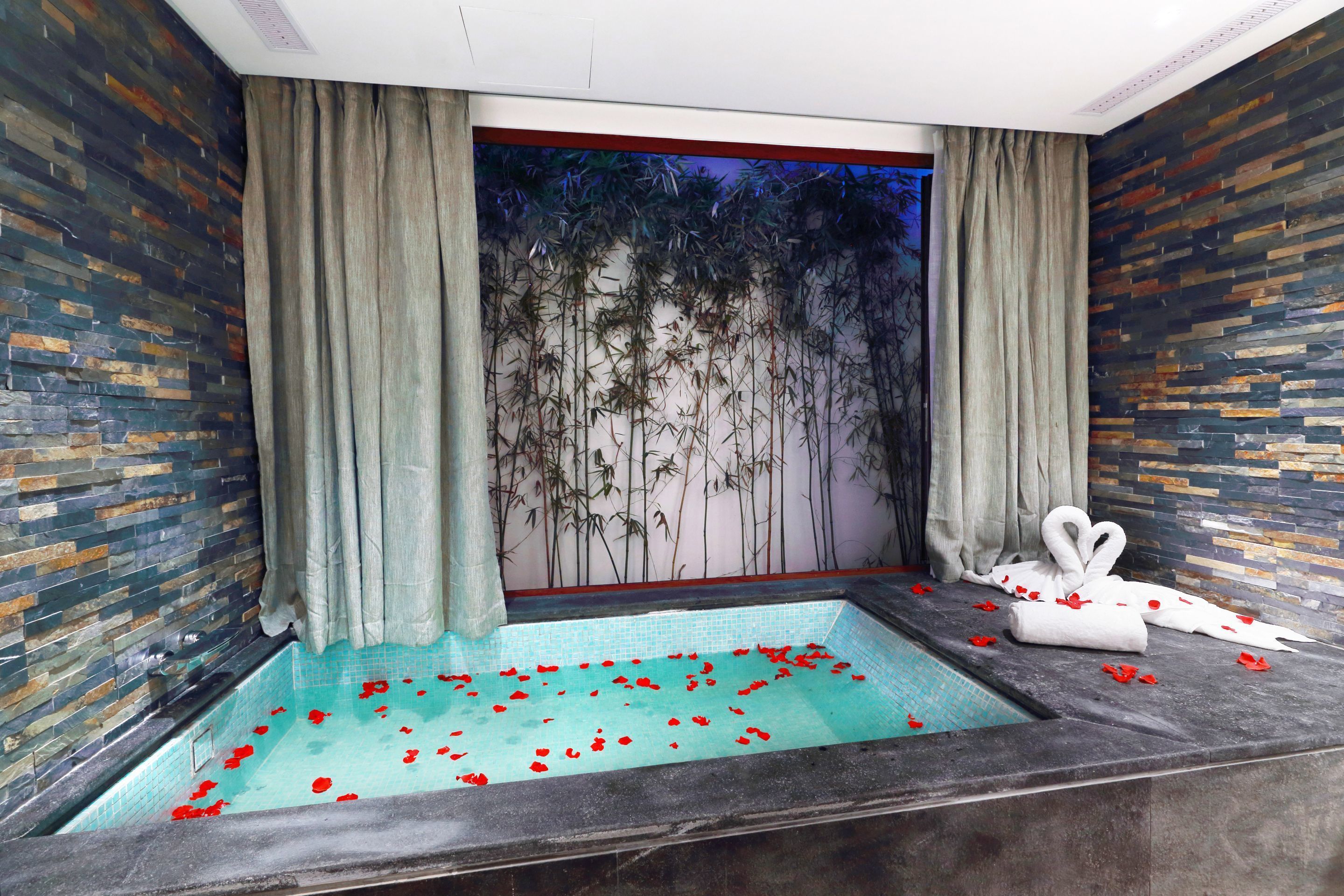 A spa room with a stone wall, a large window with curtains, a bathtub with rose petals, and a towel with a heart design.