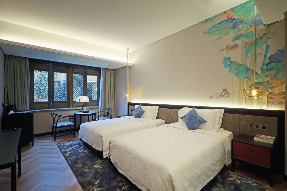 A hotel room with two beds, a desk, a chair, a window with curtains, a nightstand, and a wall mural.