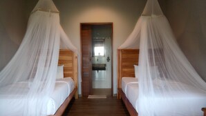 A room with two wooden beds, each with a canopy. There is a wooden bench at the foot of each bed. A door leads to a bathroom with a sink and mirror. - Emir surfcamp (Pekutatan)