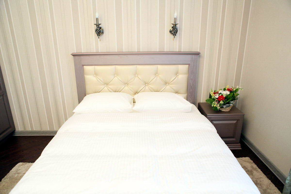 Un lit bien fait, avec une tête de lit capitonnée, dans une chambre d’hôtel aux murs recouverts d’un papier peint à rayures et une table de chevet ornée d’un vase de fleurs.