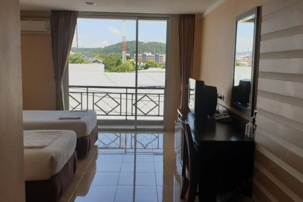 A hotel room with two beds, a TV, a desk, and a balcony with a view of the city and hills.