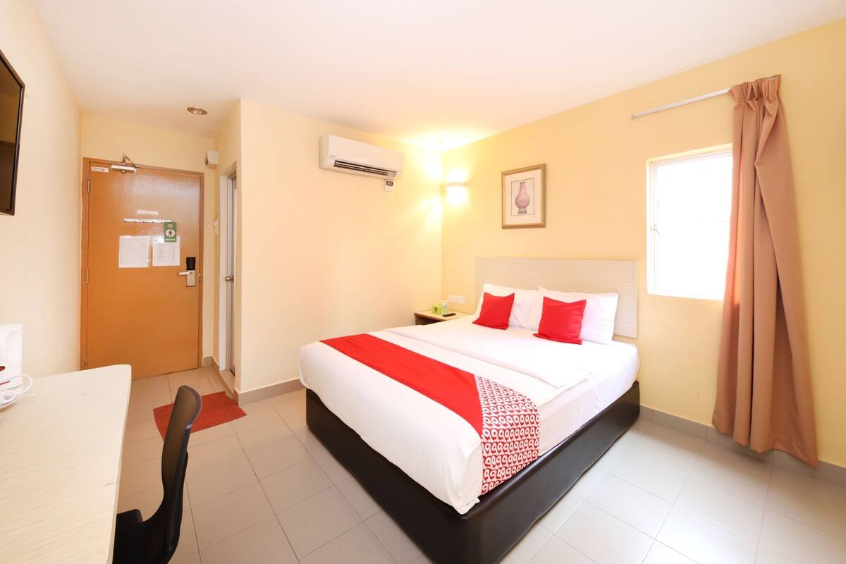 Une chambre d’hôtel comprenant un lit, un bureau, une chaise, un appareil de climatisation, une fenêtre avec des rideaux et une porte.