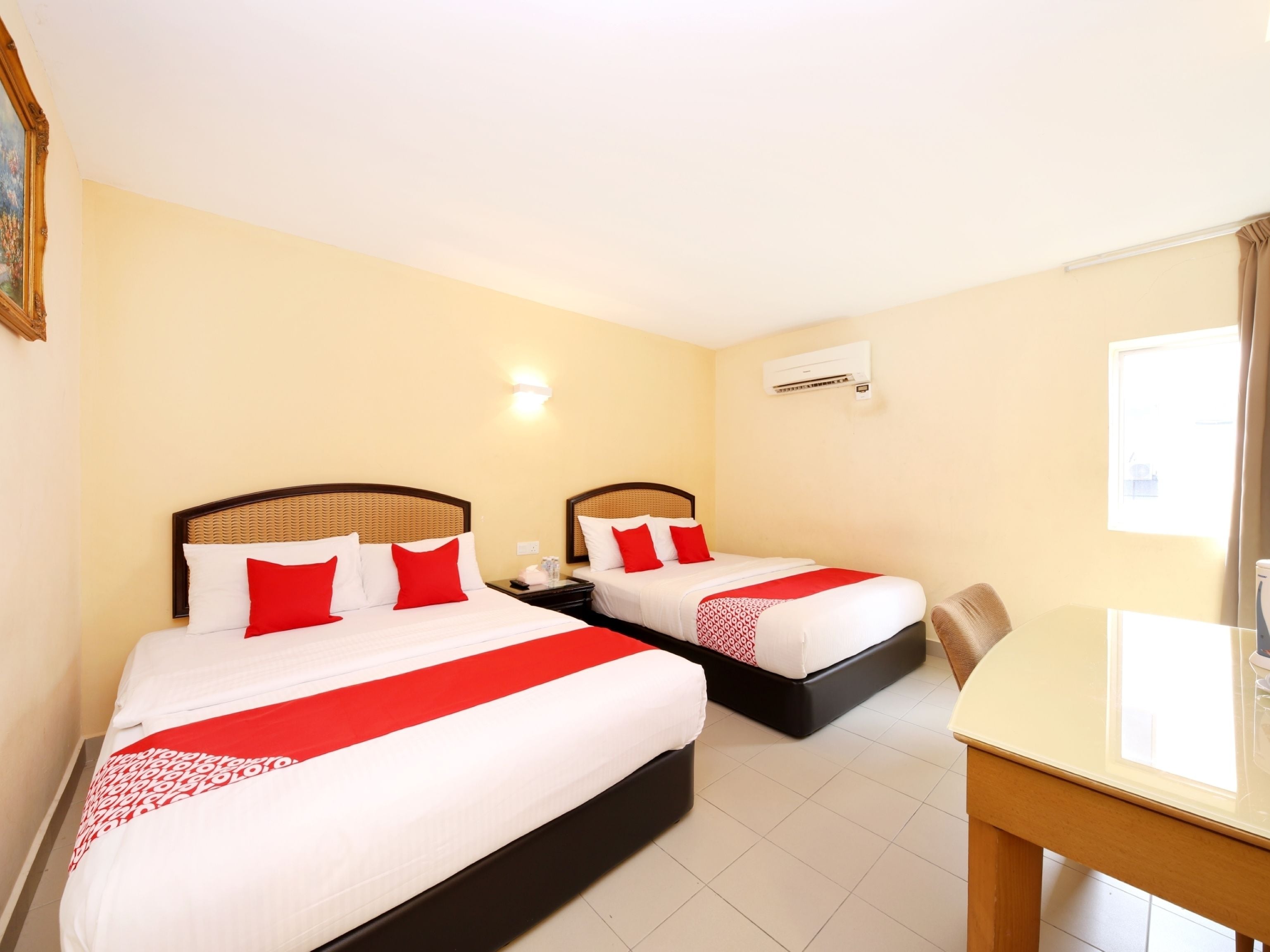 A hotel room with two beds, each with red and white bedding, a small table, and a wall-mounted air conditioner.