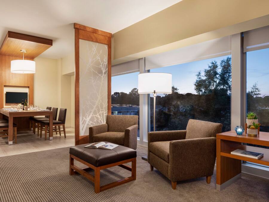 Ein modernes Hotelzimmer mit Essbereich, zwei Sessel, einem Couchtisch und großen Fenstern mit Blick auf Bäume und Gebäude.
