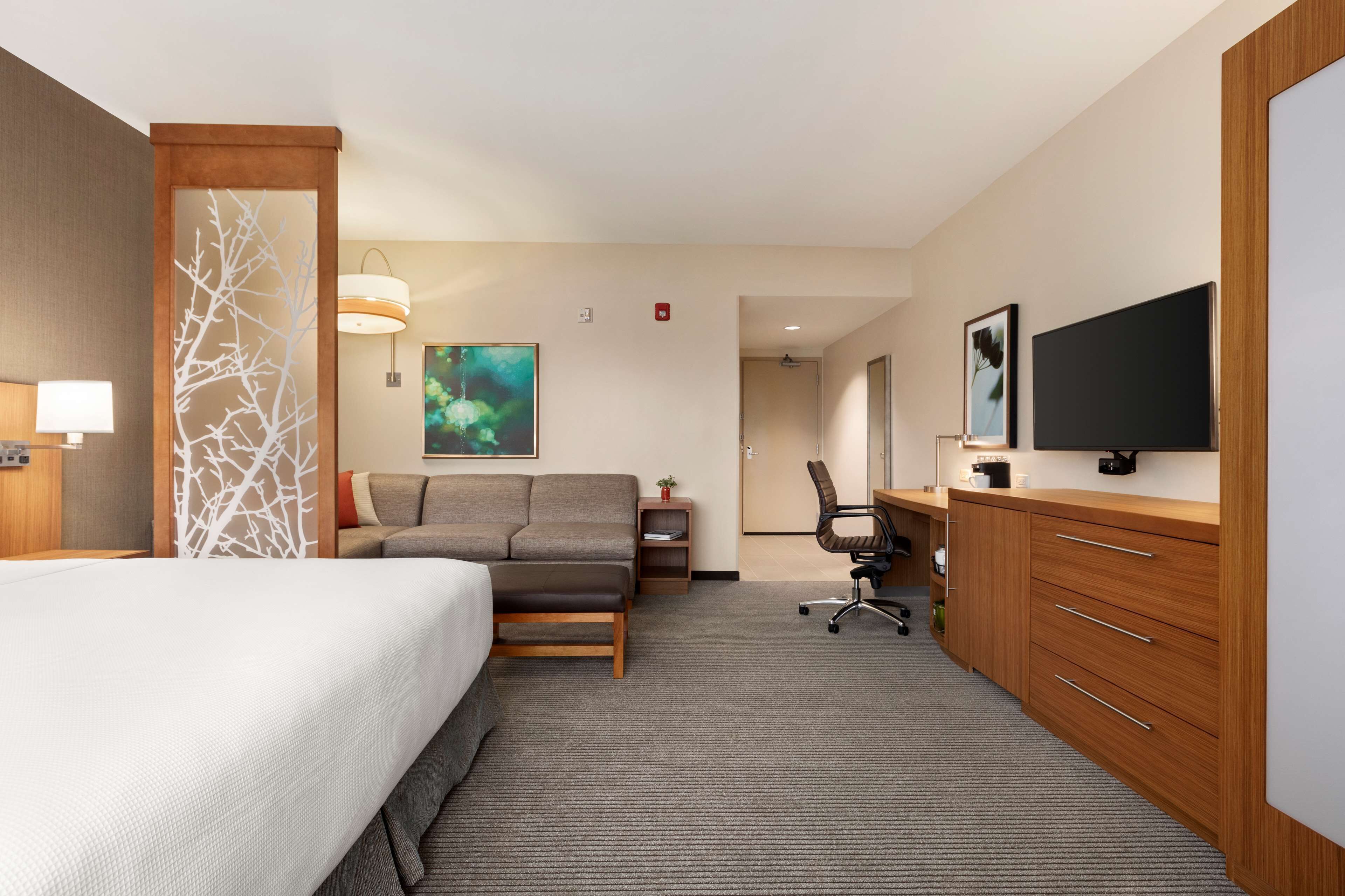 A hotel room with a bed, a sofa, a desk with a chair, a television, and a large window.
