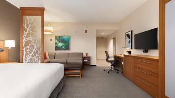 A hotel room with a bed, a sofa, a desk with a chair, a television, and a large window.
