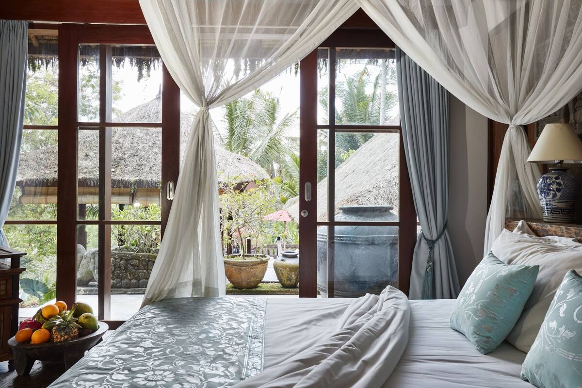 A bedroom with a bed, a bedside table with fruit, and a view of a thatched-roof structure outside.