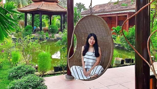 A person sitting in a wicker swing chair in a garden with a pond, a gazebo, and a traditional house in the background. - Areca Garden Bungalow (Phu Quoc)