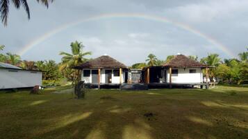 Two thatched-roof huts with white walls, surrounded by palm trees and a grassy area.