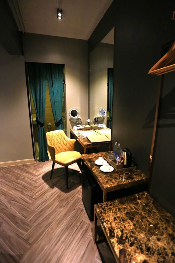 A compact hotel room with a wooden herringbone floor, a single yellow chair, a small table with a mirror, and a bed with a headboard. - Ratchada Boutique Hotel (Bangkok)