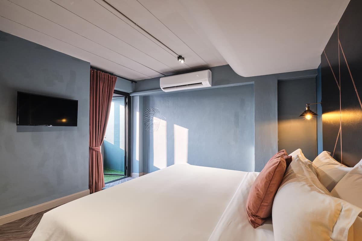 Moderni hotellihuone, jossa on suuri sänky, taulutelevisio, ilmastointilaite ja lasiovesta avautuva näkymä puutarhaan.